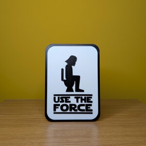 Puede incluir: Un cartel blanco con una silueta negra de Darth Vader sentado en un inodoro. El cartel dice "Use the Force" en letras negras.