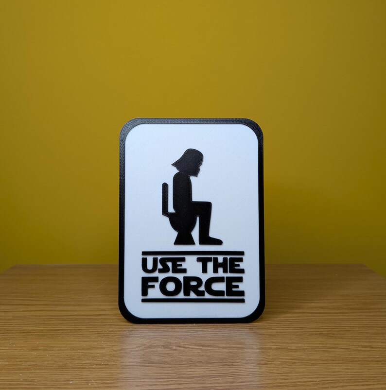 Puede incluir: Un cartel blanco con una silueta negra de Darth Vader sentado en un inodoro. El cartel dice "Use the Force" en letras negras.