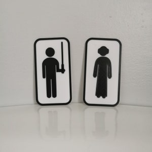 Pu&ograve; includere: Due cartelli da bagno bianchi con bordi neri. Il cartello a sinistra presenta una figura stilizzata che tiene una spada laser. Il cartello a destra presenta una figura stilizzata che indossa una tunica con cappuccio.