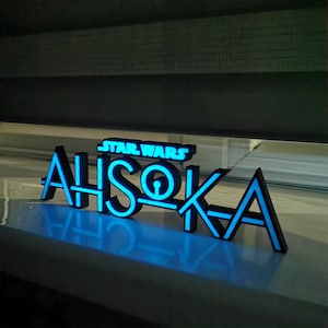 May include: A black and blue illuminated sign that reads "STAR WARS AHSOKA". The sign is made of a dark material with blue neon-style lighting outlining the letters and the Star Wars logo. The sign is placed on a white surface.