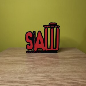May include: A red and black wooden sign with the word "SAW" in red letters and a black trash can icon. The sign is on a wooden surface.