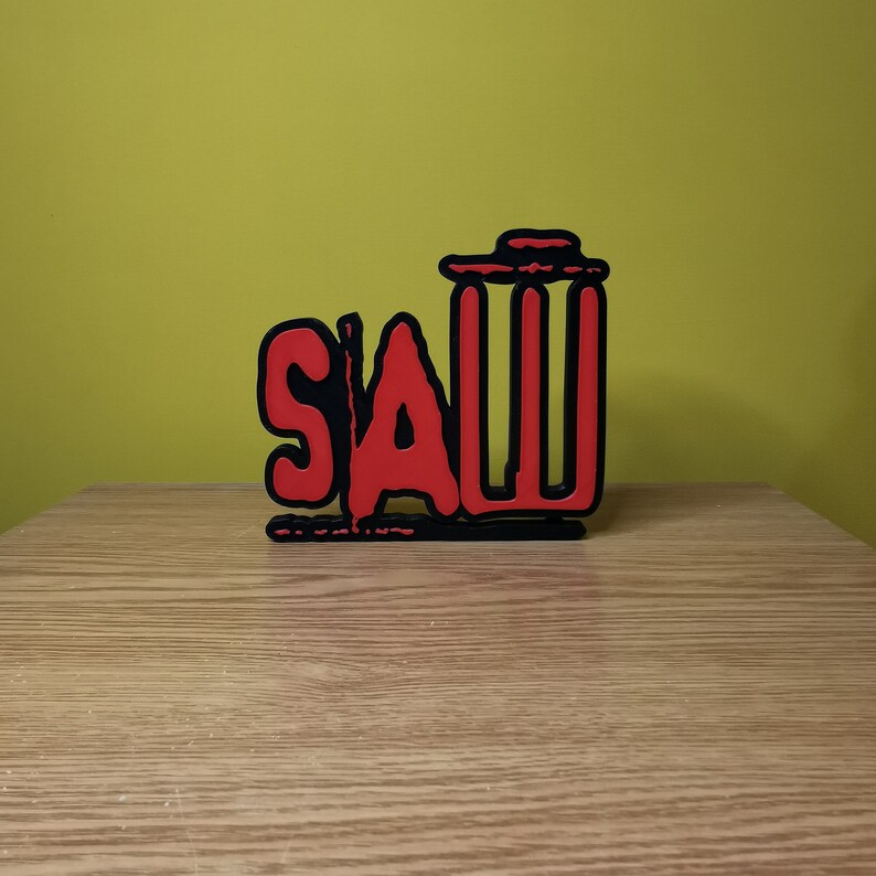 May include: A red and black wooden sign with the word "SAW" in red letters and a black trash can icon. The sign is on a wooden surface.