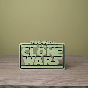 May include: A 3D printed model of the Star Wars: The Clone Wars logo. The logo is in a black frame and the text is in a light yellow color.