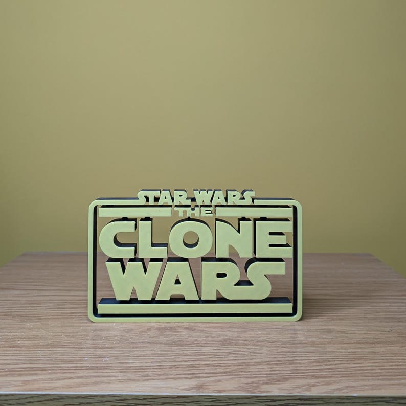 May include: A 3D printed model of the Star Wars: The Clone Wars logo. The logo is in a black frame and the text is in a light yellow color.