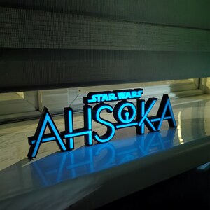 May include: A black and blue neon sign that reads "STAR WARS AHSOKA". The sign is illuminated with a bright blue light, creating a glowing effect. The sign is placed on a white surface.