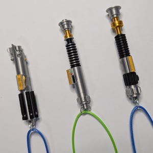 May include: Three miniature lightsaber replicas with silver, black, and gold accents. Each has a different design, with one featuring a black ribbed handle and another with a black grip. They are attached to blue and green cords.