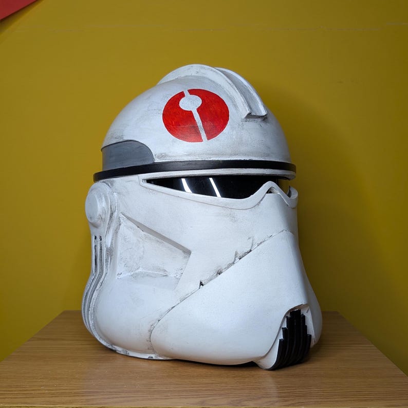 May include: A white and gray helmet with a red circular emblem. The helmet has a black visor and is designed with angular features. The helmet is likely a costume piece or collectible.