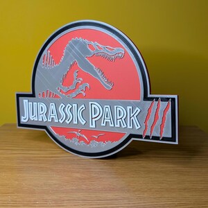 Op de afbeelding: Een 3D-geprint Jurassic Park-logo. Het heeft een rode achtergrond met een zilveren dinosaurus silhouet. De tekst "JURASSIC PARK" is wit, met een zwarte rand. Het logo bevat klauwsporen.