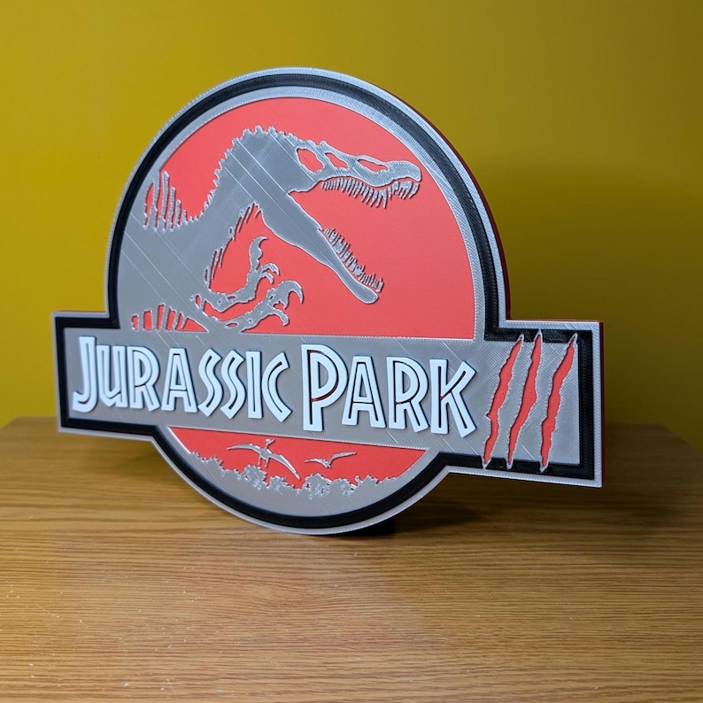 Op de afbeelding: Een 3D-geprint Jurassic Park-logo. Het heeft een rode achtergrond met een zilveren dinosaurus silhouet. De tekst "JURASSIC PARK" is wit, met een zwarte rand. Het logo bevat klauwsporen.