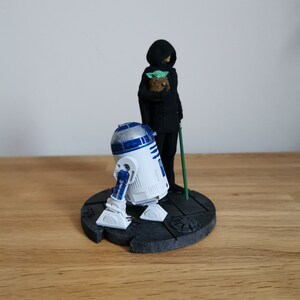 May include: A Star Wars themed figurine set featuring R2-D2, a cloaked figure holding a small green figure, and a green lightsaber. The figures are standing on a black circular base.
