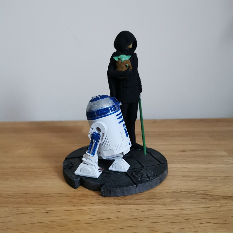 May include: A Star Wars themed figurine set featuring R2-D2, a cloaked figure holding a small green figure, and a green lightsaber. The figures are standing on a black circular base.