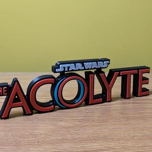 May include: A black and red 3D printed sign with the text "STAR WARS THE ACOLYTE" in white and silver lettering. The "O" in "ACOLYTE" is a blue and red circle.