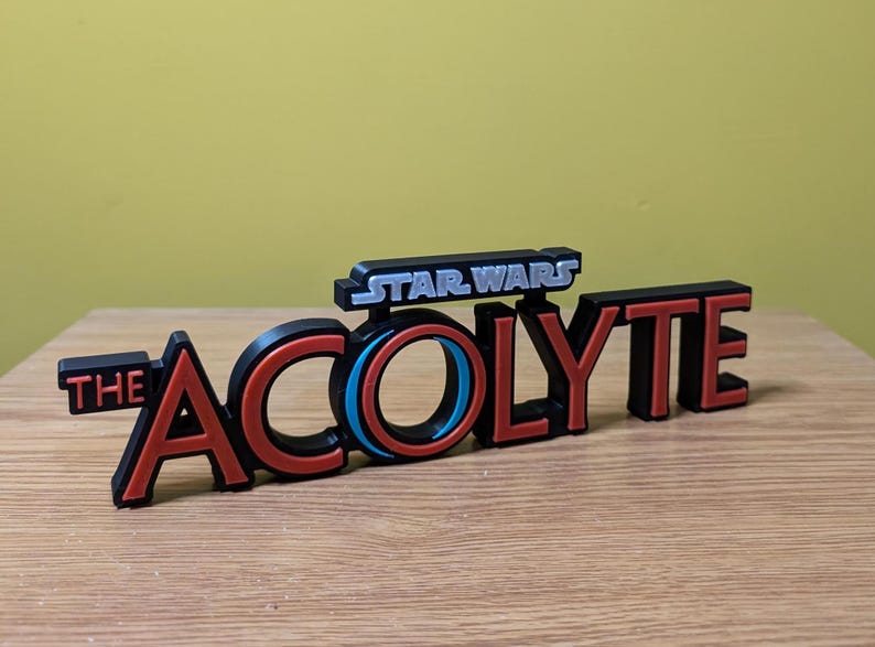 May include: A black and red 3D printed sign with the text "STAR WARS THE ACOLYTE" in white and silver lettering. The "O" in "ACOLYTE" is a blue and red circle.