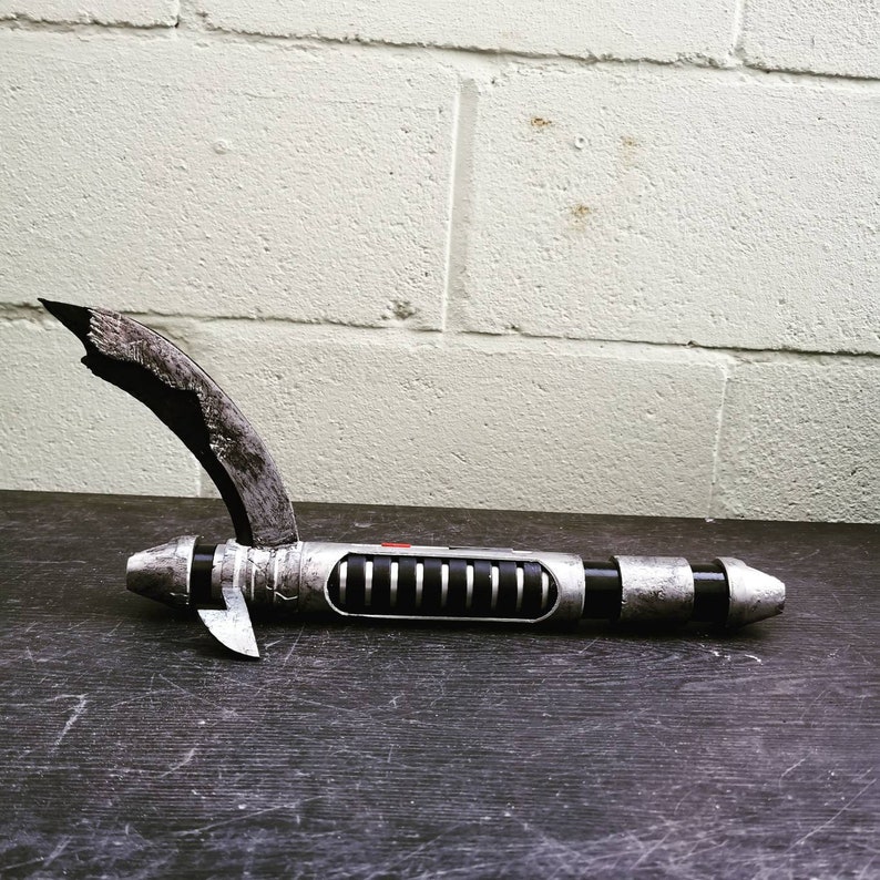 May include: A silver and black lightsaber with a broken blade, resting on a dark surface. The lightsaber has a red button and a series of black lines along the handle.