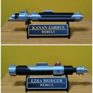 May include: Two Star Wars lightsaber replicas on display stands. The top lightsaber is silver and black with a black plaque that reads "KANAN JARRUS REBELS". The bottom lightsaber is silver and black with a black plaque that reads "EZRA BRIDGER REBELS".
