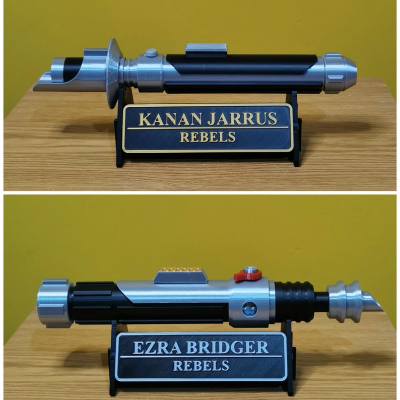 May include: Two Star Wars lightsaber replicas on display stands. The top lightsaber is silver and black with a black plaque that reads "KANAN JARRUS REBELS". The bottom lightsaber is silver and black with a black plaque that reads "EZRA BRIDGER REBELS".