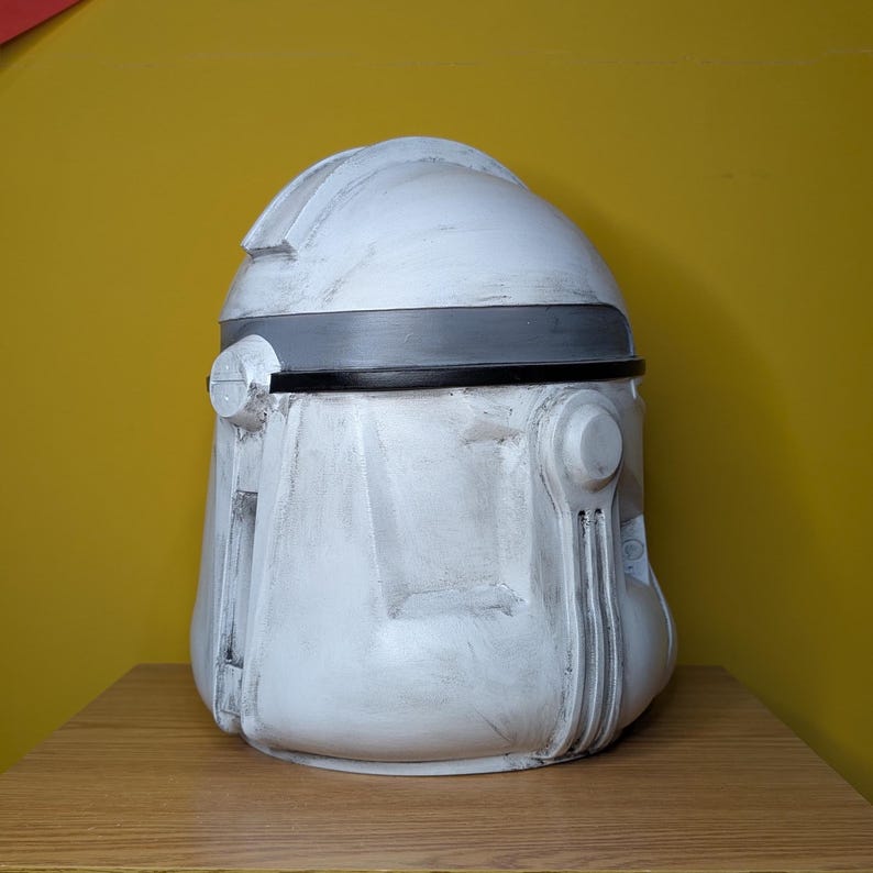 May include: A white and gray helmet with a dark gray band around the middle. The helmet has a rounded top and detailed features, suggesting a futuristic or sci-fi theme. The helmet is sitting on a wooden surface.