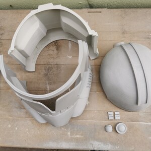 May include: A gray plastic helmet in multiple pieces, including a dome, a front piece, and two side pieces. There are also several small gray plastic pieces.