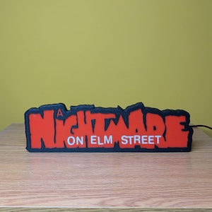 May include: A rectangular sign with the text "A NIGHTMARE ON ELM STREET" in red letters with a black outline. The sign is on a wooden surface with a yellow background.