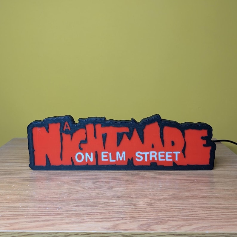 May include: A rectangular sign with the text "A NIGHTMARE ON ELM STREET" in red letters with a black outline. The sign is on a wooden surface with a yellow background.