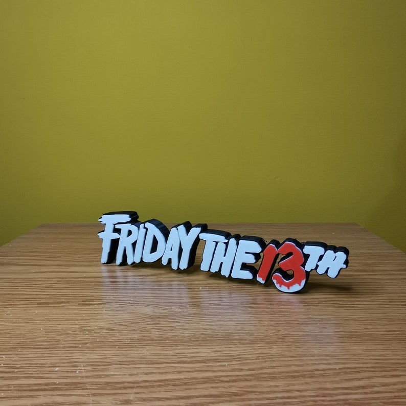 Puede incluir: Un letrero impreso en 3D en blanco y negro que dice "FRIDAY THE 13TH" con el "13" en rojo. El letrero est&aacute; sobre una superficie de madera.