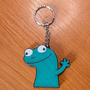 Peut inclure: Porte-cl&eacute;s en forme de l&eacute;zard de dessin anim&eacute; bleu sarcelle avec un porte-cl&eacute;s et une cha&icirc;ne argent&eacute;s. Le l&eacute;zard a de grands yeux blancs, une bouche souriante et une main lev&eacute;e. Sur une surface en bois.