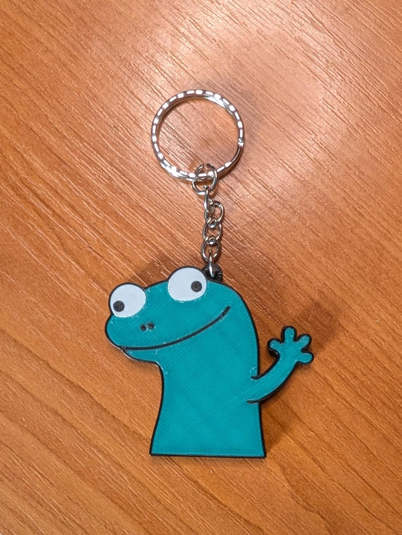 Peut inclure: Porte-cl&eacute;s en forme de l&eacute;zard de dessin anim&eacute; bleu sarcelle avec un porte-cl&eacute;s et une cha&icirc;ne argent&eacute;s. Le l&eacute;zard a de grands yeux blancs, une bouche souriante et une main lev&eacute;e. Sur une surface en bois.