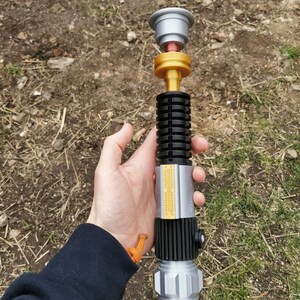 Puede incluir: Un accesorio de sable de luz plateado y dorado con un mango negro y una hoja plateada. El sable de luz tiene un agarre negro y dorado con una tapa plateada en el extremo. El sable de luz est&aacute; sostenido en la mano de una persona.