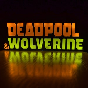Pu&ograve; includere: Insegna luminosa con la scritta "DEADPOOL & WOLVERINE" in lettere maiuscole e in grassetto. La parola "DEADPOOL" &egrave; arancione, mentre "WOLVERINE" &egrave; verde. Il riflesso dell'insegna &egrave; visibile su una superficie lucida, su sfondo scuro.