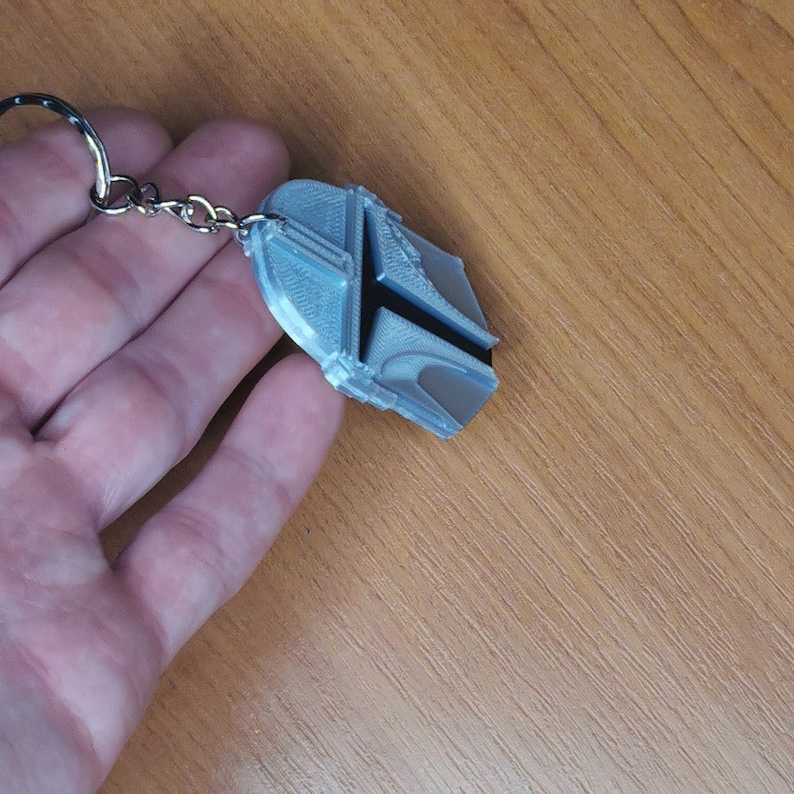 May include: A silver-colored keychain with a 3D printed design of a Mandalorian helmet. The helmet is black and silver and has a textured surface.