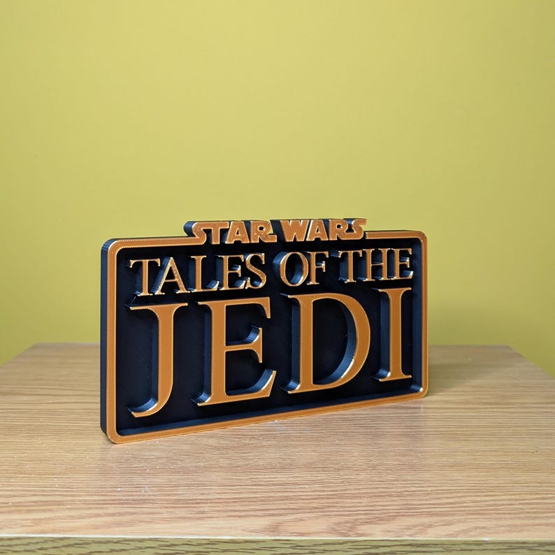 May include: A rectangular sign with the words "STAR WARS TALES OF THE JEDI" in gold lettering. The sign has a black background and a gold border. The sign is sitting on a wooden surface.