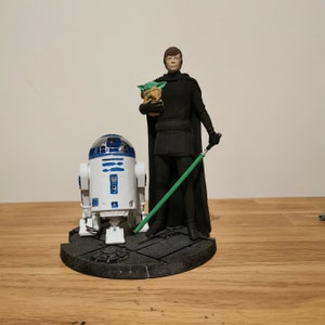 May include: A Star Wars themed figurine set featuring a black-clad figure holding a green lightsaber and a small green creature. The figure is standing on a gray base with a white and blue R2-D2 robot figurine in front of them.
