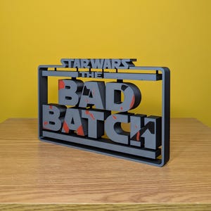 May include: A black 3D printed sign with the text "Star Wars The Bad Batch" in white letters with red accents. The sign is on a wooden surface.