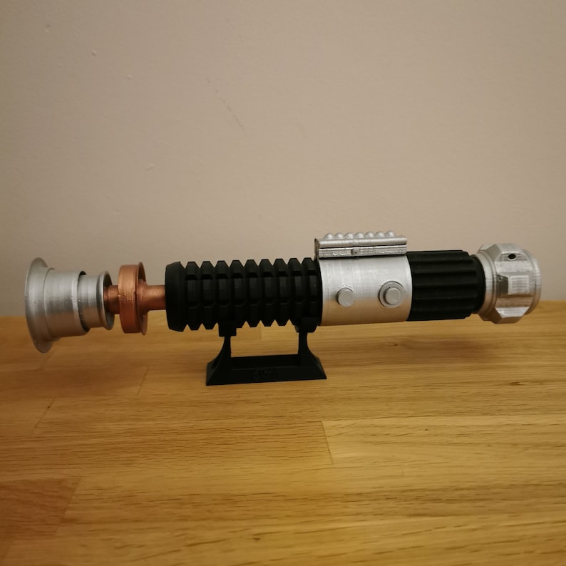 May include: A replica lightsaber prop, featuring a silver, black, and copper color scheme. The hilt has a ribbed black grip, a silver emitter, and a copper midsection. It is displayed on a black stand, set against a neutral background.