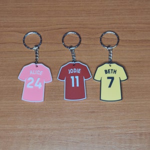Puede incluir: Tres llaveros con forma de camiseta de f&uacute;tbol. La rosa dice "ALICE 24", la roja "JODIE 11", y la amarilla "BETH 7". Cada llavero tiene un aro plateado.