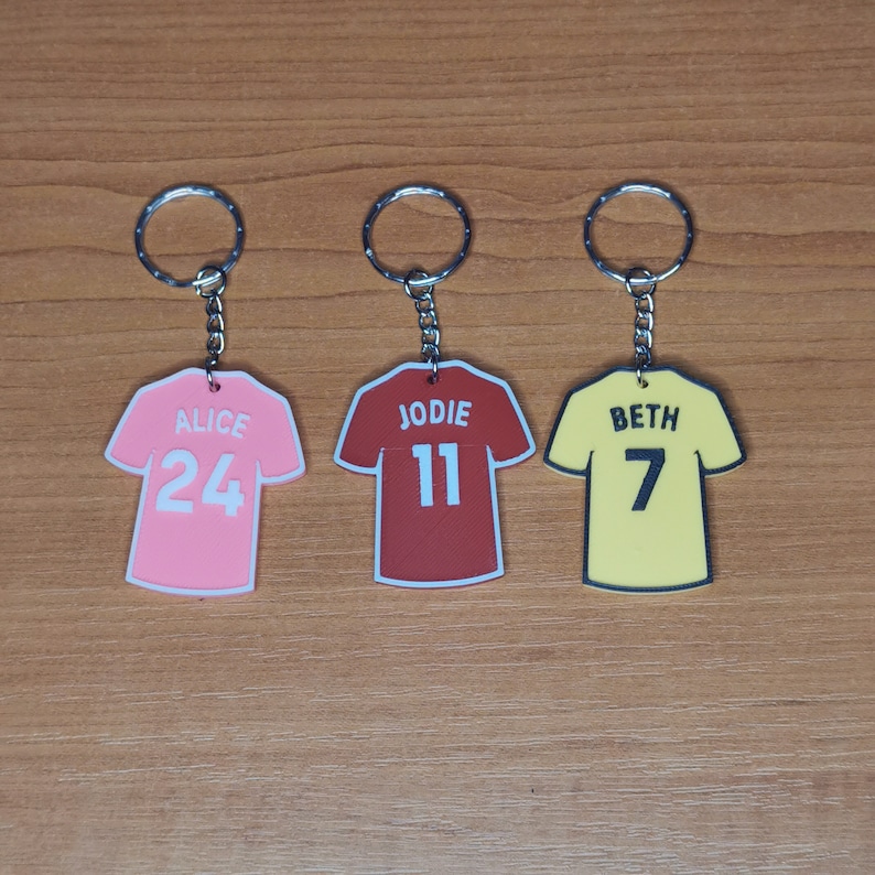 Puede incluir: Tres llaveros con forma de camiseta de f&uacute;tbol. La rosa dice "ALICE 24", la roja "JODIE 11", y la amarilla "BETH 7". Cada llavero tiene un aro plateado.