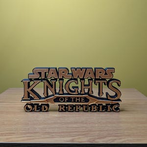 Może przedstawiać: Tr&oacute;jwymiarowy model logo gry Star Wars Knights of the Old Republic, wydrukowany w technologii 3D. Logo wykonane jest z czarnego i złotego plastiku i zawiera napis "Star Wars Knights of the Old Republic".
