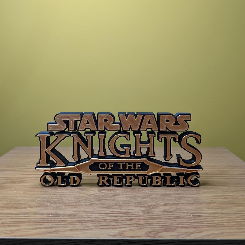 Może przedstawiać: Tr&oacute;jwymiarowy model logo gry Star Wars Knights of the Old Republic, wydrukowany w technologii 3D. Logo wykonane jest z czarnego i złotego plastiku i zawiera napis "Star Wars Knights of the Old Republic".