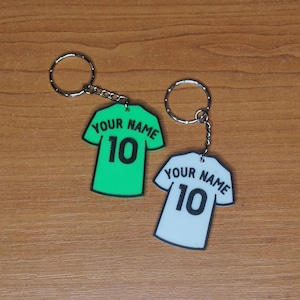 Puede incluir: Dos llaveros con forma de camisetas de f&uacute;tbol. Uno es verde brillante, el otro es blanco. Ambos tienen el texto "YOUR NAME" encima del n&uacute;mero "10" en negro. Cada llavero tiene un anillo y una cadena plateados.