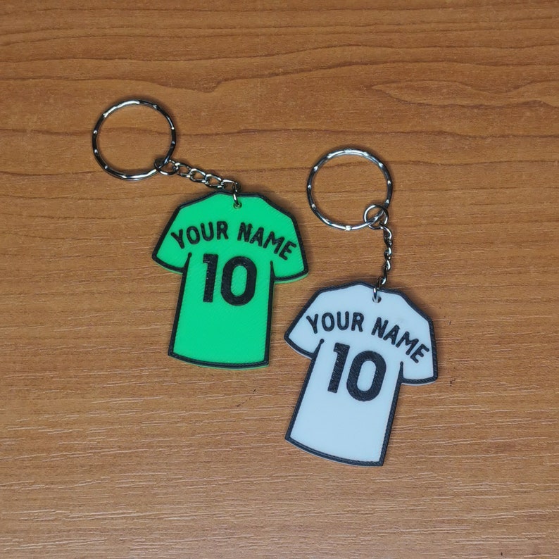 Puede incluir: Dos llaveros con forma de camisetas de f&uacute;tbol. Uno es verde brillante, el otro es blanco. Ambos tienen el texto "YOUR NAME" encima del n&uacute;mero "10" en negro. Cada llavero tiene un anillo y una cadena plateados.