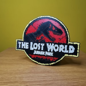 May include: A black and red 3D printed sign with the text "The Lost World Jurassic Park" in white. The sign features a silhouette of a dinosaur.