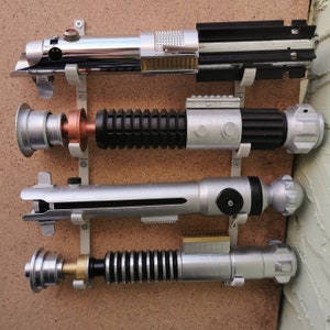 May include: Four silver and black lightsaber replicas mounted on a wall with a corkboard background. The lightsabers are arranged vertically, with the top lightsaber being the longest and the bottom lightsaber being the shortest.