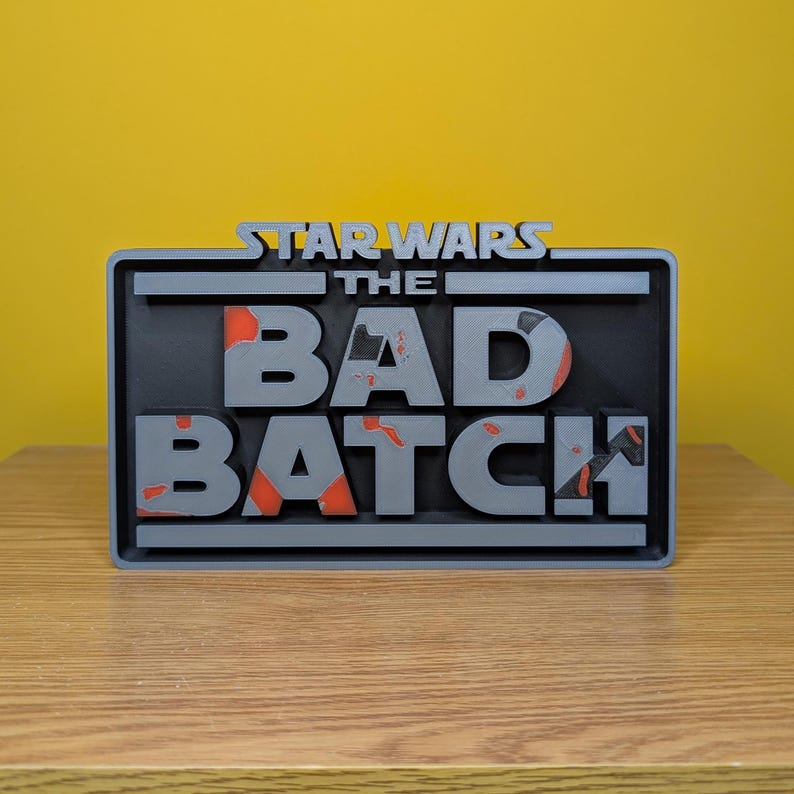 May include: A gray 3D printed sign with the text "STAR WARS THE BAD BATCH" in a distressed style. The letters are raised and have a black background with orange accents.