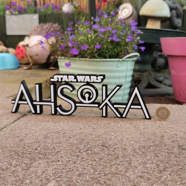 May include: Black and white Star Wars "Ahsoka" sign. The sign is made of a solid material and is placed on a concrete surface. The sign is in front of a garden with purple flowers and decorative items.