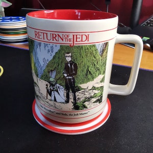 Puede incluir: Una taza de cer&aacute;mica blanca con un borde y un asa rojos. La taza presenta una ilustraci&oacute;n de estilo c&oacute;mic de Luke Skywalker, Yoda y una escena de bosque de la pel&iacute;cula El Retorno del Jedi. La taza est&aacute; sentada sobre un posavasos blanco con un borde rojo.