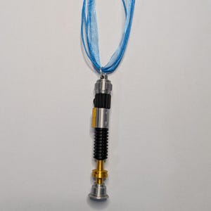 May include: A silver, black, and gold lightsaber pendant hangs from a light blue ribbon necklace. The pendant has a detailed design with a cylindrical body, a black ribbed section, and a gold-colored base. The ribbon is made of multiple strands.