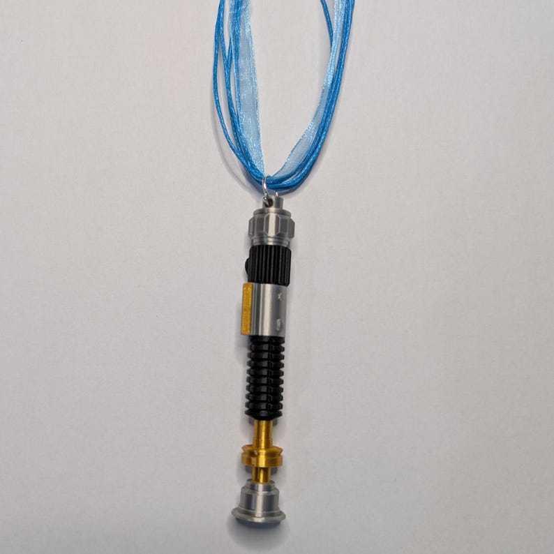 May include: A silver, black, and gold lightsaber pendant hangs from a light blue ribbon necklace. The pendant has a detailed design with a cylindrical body, a black ribbed section, and a gold-colored base. The ribbon is made of multiple strands.