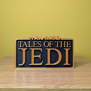 May include: A rectangular sign with the words "STAR WARS TALES OF THE JEDI" in gold lettering on a navy blue background. The sign has a gold border and is resting on a wooden surface.