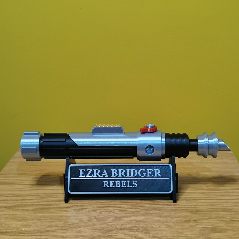 May include: A silver and black lightsaber with a red button on the side, on a black stand with a plaque that reads "Ezra Bridger Rebels".