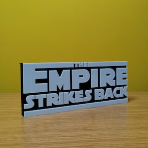 May include: A black and white sign with the text "THE EMPIRE STRIKES BACK" in white raised letters on a black background.